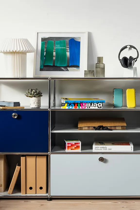 Modernes USM Chromregal mit blauem und grauem Fach, dekoriert mit Büchern und Lampe. Daneben steht eine Akustikgitarre.