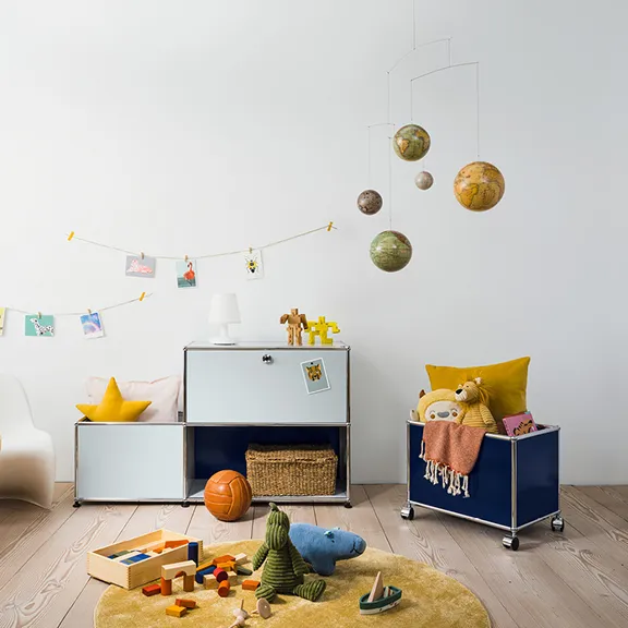 Helles Kinderzimmer mit weiß-blauem USM Chromregal, passendem Rollcontainer und Spielzeug auf einem runden gelben Teppich.